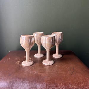 Puede incluir: Cuatro copas de vino de cerámica con un diseño texturizado y acanalado. Las copas tienen un color beige claro con sutiles acentos iridiscentes. Los tallos son delgados y las bases son redondas. Las copas están dispuestas sobre una superficie marrón sobre un fondo verde oscuro.