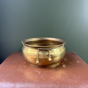 May include: A round, polished brass bowl featuring a decorative twisted rope trim encircling the upper rim, with two short, dangling rope-like accents ending in small spherical beads hanging from the front of the metallic vessel.