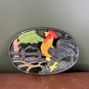 Peut inclure: Décoration murale ovale en fonte représentant un coq. Le coq est peint avec des détails rouges, oranges, jaunes et noirs. Le motif comprend un arbre, une clôture et du feuillage. La bordure est noire.