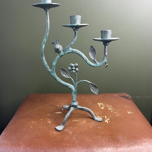 Puede incluir: Un candelabro de metal de tres brazos con acabado en color verde verdigris, que presenta una base de trípode, un motivo floral central y una pequeña figura de pájaro posada en una de las ramas curvas. Cada brazo sostiene un portavelas circular.