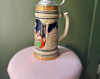 May include: A ceramic beer stein with a detailed relief scene, featuring figures in traditional attire. The stein has a silver-coloured metal lid and handle. The colours include blue, beige, brown, and touches of red and green. It is sitting on a small stool.