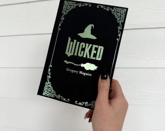 May include: A black book with the title "Wicked" in a stylized font, featuring a witch's hat and broom illustration. The book cover has decorative corner designs and the author's name, Gregory Maguire, is printed below the title.