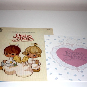 May include: Two "Precious Moments" items. One is a cross-stitch pattern featuring two figures with halos and wings. The other is a card with a pink heart and the words "Precious Moments".
