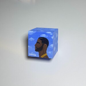 May include: A hand-painted wooden cube featuring a portrait of Drake in profile. The cube is painted with a blue sky background and white clouds. The word "DRAKE" is written on the side of the cube.