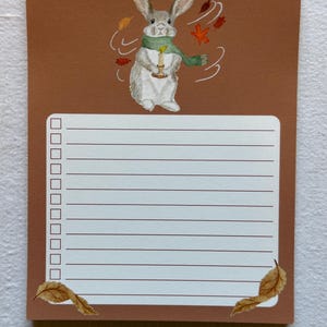 May include: A brown notepad with a watercolor illustration of a gray rabbit wearing a green scarf and holding a candle. The notepad has a lined section for notes and check boxes. Autumn leaves are scattered around the rabbit and at the bottom corners.