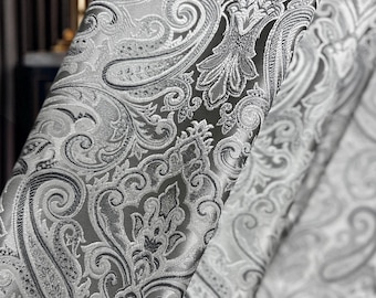 May include: A curtain fabric featuring a dense, intricate paisley and damask pattern in shades of silver, charcoal gray, and white. The design consists of swirling teardrop shapes and ornate floral motifs woven into a textured, reflective material.