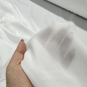 May include: Close-up of a hand holding a piece of sheer white fabric. The fabric is lightweight and slightly textured, with visible light passing through. The background shows more of the same white fabric, possibly a roll or a larger piece.