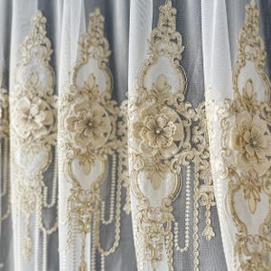 Puede incluir: Primer plano de cortinas blancas transparentes con intrincados bordados florales y de volutas en crema y dorado. El diseño presenta aplicaciones florales en 3D y detalles de cuentas, creando un aspecto elegante y texturizado. Las cortinas están parcialmente cerradas.