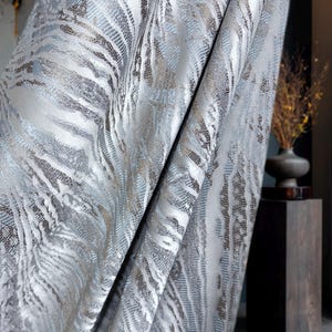 May include: A heavy fabric curtain featuring a textured, abstract pattern with vertical waves in shades of silver, metallic bronze, and light blue. The material displays a dense, woven finish with a subtle sheen, creating deep folds and shadows.