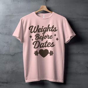 May include: A light pink t-shirt with the phrase "Weights Before Dates" in dark brown lettering. The design includes a heart and barbell graphic. The shirt is hanging on a wooden hanger.