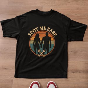 以下が含まれることがあります： レトロなデザインの黒いTシャツ。円形のフレームの中で手をつないでいる2つのシルエットが特徴です。「SPOT ME BABE」の文字が人物の上にアーチ状に配置されています。ダンベルが円の下部にあります。
