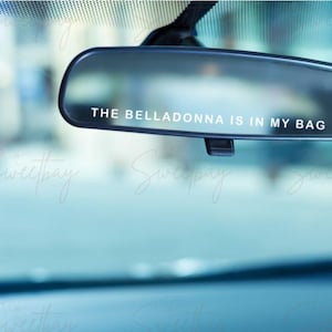 Op de afbeelding: Een zwarte achteruitkijkspiegel met de witte tekst "THE BELLADONNA IS IN MY BAG". De achtergrond is wazig, wat duidt op een straatbeeld. De afbeelding heeft een koele, enigszins raadselachtige uitstraling.