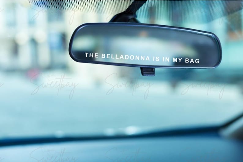 Op de afbeelding: Een zwarte achteruitkijkspiegel met de witte tekst "THE BELLADONNA IS IN MY BAG". De achtergrond is wazig, wat duidt op een straatbeeld. De afbeelding heeft een koele, enigszins raadselachtige uitstraling.