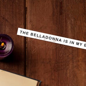 Op de afbeelding: Een brandende paarse kaars met een gele vlam staat op een donker houten oppervlak. Een wit rechthoekig bord met de tekst "THE BELLADONNA IS IN MY BAG" is op het hout geplaatst. Een boek is gedeeltelijk zichtbaar.