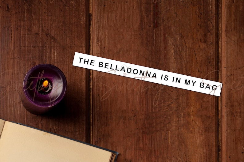 Op de afbeelding: Een brandende paarse kaars met een gele vlam staat op een donker houten oppervlak. Een wit rechthoekig bord met de tekst "THE BELLADONNA IS IN MY BAG" is op het hout geplaatst. Een boek is gedeeltelijk zichtbaar.