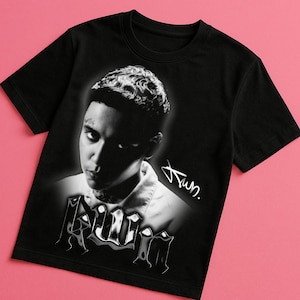 May include: Black t-shirt featuring a grayscale portrait of a person with a tattoo and signature. The word "Killin" is printed in a stylized font at the bottom of the portrait. The shirt is displayed on a pink background.