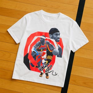 Op de afbeelding: Wit T-shirt met een afbeelding van een basketballer in een rood en blauw uniform, met een basketbal en een handtekening. Het ontwerp is geplaatst tegen een rode en zwarte geometrische achtergrond. Het shirt heeft een ronde hals.
