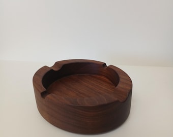 May include: A round, wooden ashtray with a dark brown finish. The ashtray has a low profile with curved edges and three small grooves for resting cigarettes. The wood grain is visible, adding texture to the piece.