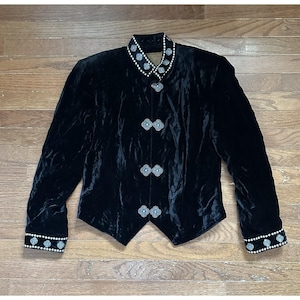 May include: A black velvet jacket featuring a high collar and long sleeves with white beaded trim and silver-toned floral-shaped metal accents. The front closure consists of three matching silver-toned metal clasps, creating a structured silhouette.