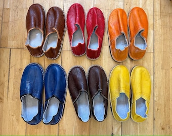 May include: A collection of leather slippers in various colors, including brown, red, orange, blue, dark brown, and yellow. The slippers are arranged in rows on a wooden surface, showcasing their different hues and designs.