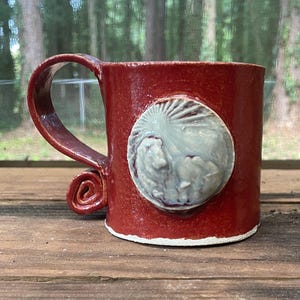 May include: A handmade ceramic mug with a deep red glaze. The mug features a sculpted, circular design in a lighter color, possibly depicting a sun or celestial body. The handle has a unique spiral detail. The mug is approximately 4 inches tall.