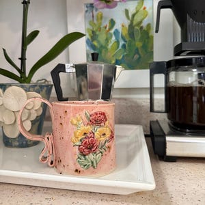 May include: A pink ceramic mug with a speckled finish and a sculpted floral relief featuring red and yellow flowers with green leaves. The mug has a unique, looped handle design and sits on a white rectangular tray.