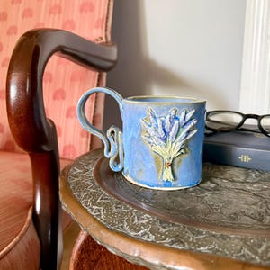 May include: A light blue ceramic mug featuring a raised, sculpted lavender bouquet on the front. The mug has a unique, curved handle with a decorative loop design. The glaze has subtle variations in tone, highlighting the textured floral relief.