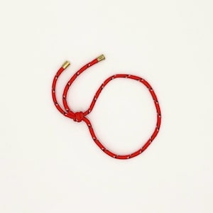 May include: A red and white rope bracelet with gold-colored metal tips. The bracelet is knotted in the center and has a circular shape. The rope has a textured appearance and is set against a white background.