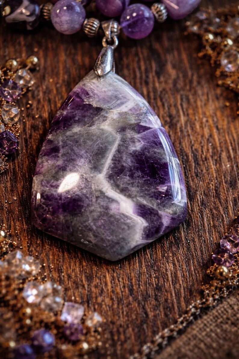 Puede incluir: Un colgante grande de amatista pulida con una fianza de plata. El colgante es de color p&uacute;rpura oscuro con vetas grises y blancas m&aacute;s claras. El fondo incluye una superficie de madera y otras cuentas de amatista.