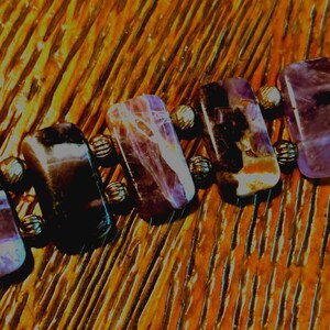Puede incluir: Primer plano de una pulsera con piedras preciosas rectangulares pulidas en tonos morados y marrones. Las piedras est&aacute;n conectadas por peque&ntilde;as cuentas met&aacute;licas redondas. La pulsera est&aacute; sobre una superficie de madera.