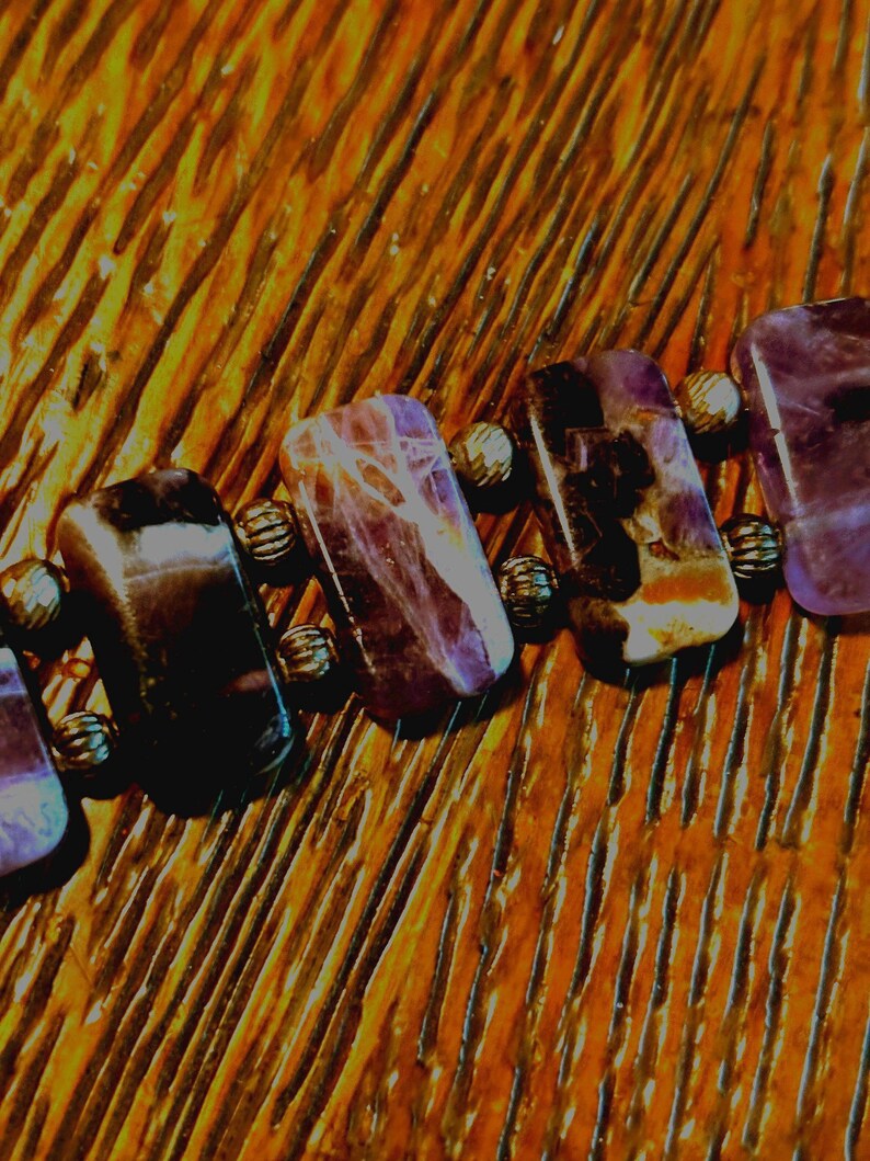 Puede incluir: Primer plano de una pulsera con piedras preciosas rectangulares pulidas en tonos morados y marrones. Las piedras est&aacute;n conectadas por peque&ntilde;as cuentas met&aacute;licas redondas. La pulsera est&aacute; sobre una superficie de madera.