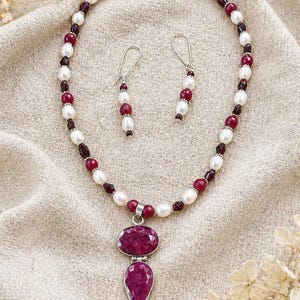 May include: A necklace and earring set with a silver pendant holding two ruby stones, oval and teardrop-shaped. The necklace and earrings are strung with white pearls, red beads, and dark red garnet stones. The jewellery set is displayed on a beige fabric.