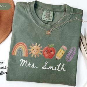 May include: A moss green short-sleeve t-shirt featuring a faux patchwork graphic of a rainbow, a smiling sun, a smiling apple, a pencil, and a paperclip. The text "Mrs. Smith" is printed in white script below the graphics. The shirt has a crew neck.
