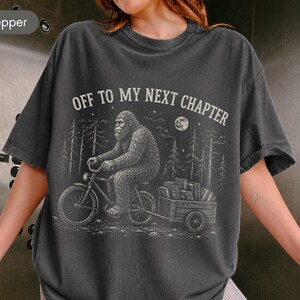 May include: A charcoal gray short-sleeve t-shirt featuring a white graphic print of a Bigfoot creature riding a bicycle and pulling a small trailer filled with books. The text above the illustration reads, OFF TO MY NEXT CHAPTER.