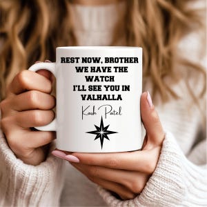 Könnte beinhalten: Weiße Keramiktasse mit schwarzem Text: "REST NOW, BROTHER WE HAVE THE WATCH I'LL SEE YOU IN VALHALLA Kash Patel" und einem schwarzen Sternmuster. Die Tasse wird von einer Person gehalten.