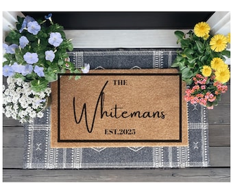 May include: A rectangular doormat with the text "The Whitemans EST.2025" in a black frame. The mat is brown and sits on a patterned gray rug. Potted flowers in shades of blue, white, yellow, and pink surround the mat.