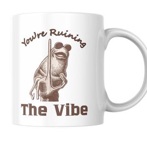 May include: White ceramic mug featuring a frog wearing sunglasses and a raised finger, with the text "You're Ruining The Vibe" in brown lettering. The mug has a curved handle for easy gripping.