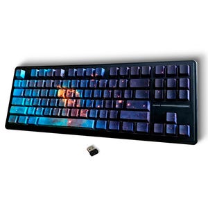 May include: A black wireless computer keyboard featuring a space-themed design on the keycaps with blue, purple, and orange nebula patterns. The keyboard includes a small black USB receiver positioned below the main unit on a plain white background.