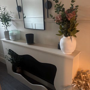 May include: A white console table with a distinctive cut-out design, displaying a small potted plant and a vase of flowers. A black arched mirror is mounted above, flanked by wall sconces. The room is decorated with plants and a vase of dried flowers.