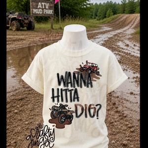 以下が含まれることがあります： 「WANNA HITTA DIG？」の文字とATVと泥しぶきのイラストが描かれたクリーム色のTシャツ。泥だらけのATVパークを背景にマネキンに着せられています。
