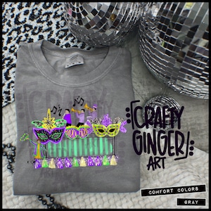 May include: Grey t-shirt with a Mardi Gras design featuring masks, musicians, and the text "Mardi Gras." The shirt also has the text "Crafty Ginger Art" and "Comfort Colors Gray."