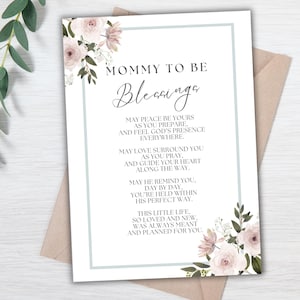 May include: A white card with the text "MOMMY TO BE Blessings" in elegant script. The card features a light blue border and is adorned with floral arrangements in shades of pink and green. The card is placed on a neutral-toned envelope.
