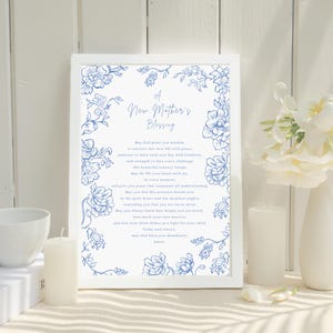 May include: A framed print featuring the text "A New Mother's Blessing" and blue floral designs. The print is set against a white background, with a white candle and a vase of white flowers, creating a serene and delicate aesthetic.