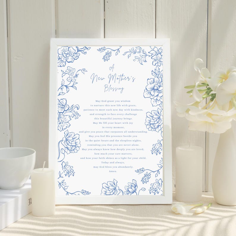 May include: A framed print featuring the text "A New Mother's Blessing" and blue floral designs. The print is set against a white background, with a white candle and a vase of white flowers, creating a serene and delicate aesthetic.