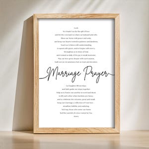 Puede incluir: Una impresión enmarcada con un marco de veta de madera clara. La impresión presenta un poema titulado "Marriage Prayer" en una escritura elegante. El texto es negro sobre un fondo blanco, creando una estética clásica y atemporal. El marco es rectangular.