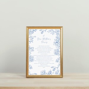 May include: A framed print with a gold-coloured frame. The print features the text "A New Mother's Blessing" in elegant script, surrounded by blue floral illustrations on a white background. The print is displayed on a light-coloured wooden surface.