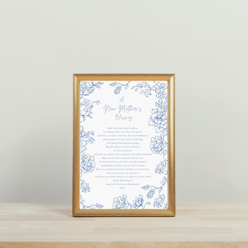May include: A framed print with a gold-coloured frame. The print features the text "A New Mother's Blessing" in elegant script, surrounded by blue floral illustrations on a white background. The print is displayed on a light-coloured wooden surface.