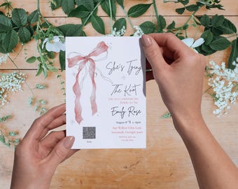 May include: A wedding invitation with the text "She's Tying The Knot" and the name "Emily Rose". The invitation features a watercolor bow design in shades of pink and is held against a wooden background with floral accents.