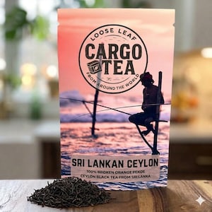 Puede incluir: Un paquete de té Cargo, etiquetado Sri Lankan Ceylon. El envase presenta un logotipo circular con las palabras "Cargo Tea" y "Loose Leaf", y una ilustración de un pescador. Un montón de hojas de té sueltas está delante del paquete.