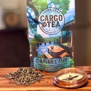 May include: A package of "Cargo Tea" loose leaf tea with the text "Chun Mee Jade" and a compass on a wooden surface. The tea is from the Jiangxi Province of China. A pile of loose green tea leaves is next to the package.